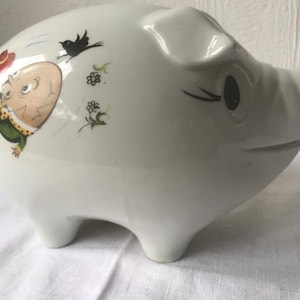 Puede incluir: Huchita de cerámica blanca con una ilustración de dibujos animados de un hombre con un traje verde y un sombrero rojo cayendo del cielo. La hucha tiene un pájaro negro y flores pintadas en ella.