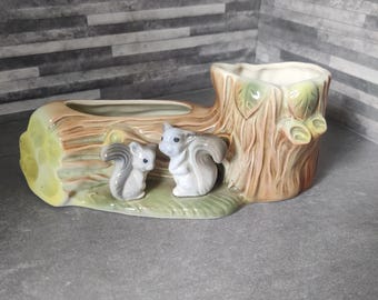 May include: A ceramic planter shaped like a log and tree stump, with two gray squirrels. The planter has a brown, tan, and green color scheme. The log section has a long opening, and the tree stump has a round opening.