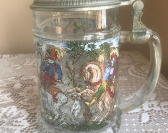 May include: A clear glass beer stein with a hinged metal lid and handle. The stein features a colourful illustration of people on horseback in a forest scene. The handle and lid are silver-toned.