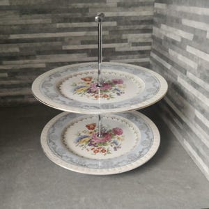 May include: Two-tiered cake stand with a silver metal stand and two white ceramic plates with a floral design. The plates have a blue and gold trim.