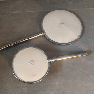 Könnte beinhalten: Zwei Handspiegel mit silbernen Griffen und weißen, runden, reflektierenden Oberflächen. Jeder Spiegel hat ein kleines, dekoratives, sternförmiges Design in der Mitte.