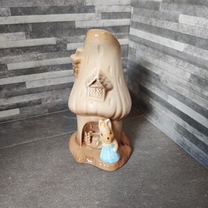 Puede incluir: Una figurita de cerámica de un conejo con un vestido azul de pie frente a una casa de hongo. La casa tiene un techo marrón y una pequeña ventana.