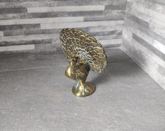 Puede incluir: Pequeña figura de pavo real de latón con una cola detallada en forma de abanico. El pavo real está de pie sobre una base redonda. La figura tiene un aspecto envejecido y antiguo, con una mezcla de tonos dorados y oscuros.