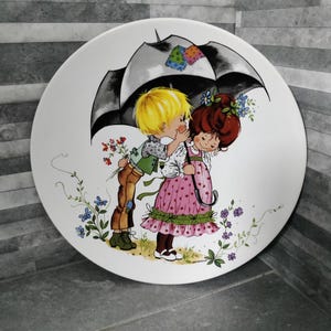 Può includere: Un piatto in ceramica bianca con un'illustrazione colorata di due bambini sotto un ombrello. La ragazza indossa un vestito rosa e il ragazzo indossa una camicia verde e pantaloni marroni. L'illustrazione è circondata da fiori.