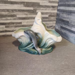 May include: A ceramic planter shaped like a wave with a dolphin jumping out of it. The planter is green, white, and grey.