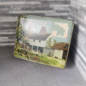 May include: Vintage tin box with a colorful image of a white farmhouse with a barn and a pond in the foreground. The tin has a gold trim around the edges.