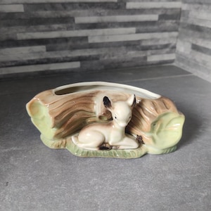 May include: A ceramic planter shaped like a log, featuring a fawn resting on grass. The planter is painted in brown, green, and white. The fawn has black ears and brown spots. Designed for small plants, it measures approximately 20 cm in length.