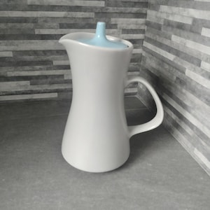 May include: White ceramic jug with a light blue lid. The jug has a wide base and a narrow neck. The lid has a small spout.