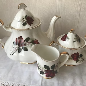 Puede incluir: Un juego de té blanco con un diseño floral con rosas rojas y hojas verdes. El juego incluye una tetera, una jarra de crema y un azucarero.