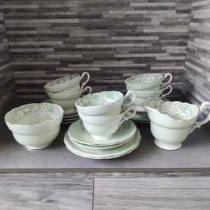 Könnte beinhalten: Ein Vintage-Teeservice in hellem Grün, bestehend aus Tassen, Untertassen und einem Sahnekännchen. Die Tassen und das Sahnekännchen haben ein zartes Blumenmuster im Inneren. Das Set umfasst mehrere Tassen, Untertassen und ein Sahnekännchen.