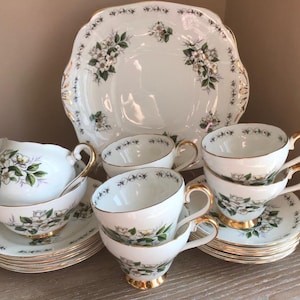 Może przedstawiać: Zestaw do herbaty w stylu vintage z kwiatowym wzorem. Zestaw zawiera czajnik, dzbanek na śmietanę, cukiernicę, cztery filiżanki do herbaty i spodki oraz talerz na ciasto. Zestaw jest biały z pozłacanymi zdobieniami i ma delikatny kwiatowy wzór.