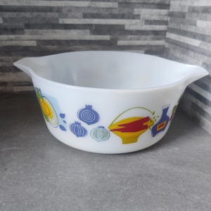 May include: White Pyrex mixing bowl with a colourful vegetable design. The bowl features illustrations of onions, lemons, and a basket of tomatoes. The bowl has two handles and is set against a grey background.