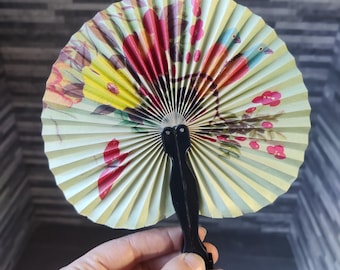 Puede incluir: Un abanico de mano con un diseño floral en tonos rojos, amarillos y verdes. El abanico está hecho de papel y tiene un mango de plástico negro.
