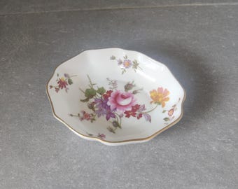May include: A white saucer with a scalloped edge and a floral design featuring pink, purple, and yellow flowers. The saucer has a gold trim around the edge.