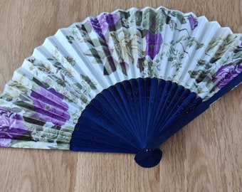 Puede incluir: Un abanico plegable blanco con un patrón floral en tonos de morado y verde. El abanico tiene un mango de madera azul con un diseño decorativo.