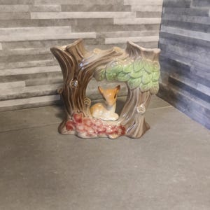 May include: A ceramic vase shaped like a tree trunk with a deer peeking out from behind the trunk. The vase is painted in shades of brown, green, and red.