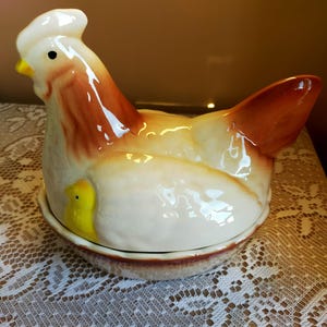 May include: Cream and brown ceramic hen-shaped container with a chick detail. The hen has brown and orange accents. The container sits on a round, brown-rimmed base. A decorative kitchen item.