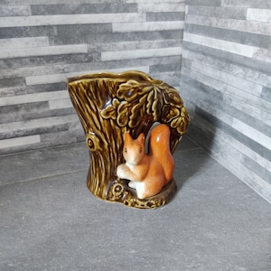 May include: A ceramic vase in the shape of a tree trunk, featuring a squirrel figurine. The vase is brown with oak leaf detailing and a glossy finish. The squirrel is orange and white, a charming piece of nature-inspired decor.