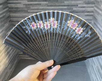 Puede incluir: Un abanico negro con un diseño floral. El abanico presenta flores rosas, amarillas y moradas con hojas verdes. Los bordes del abanico están adornados con oro. El abanico está abierto.