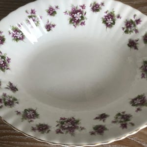 May include: A white ceramic bowl with a scalloped edge and a floral design featuring purple and white flowers. The bowl is decorated with a delicate pattern of small flowers.