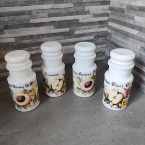 May include: Four white ceramic spice jars with lids and colorful fruit illustrations. The jars are labeled "Nutmeg", "Cinnamon", "Saffron", and "Clove".