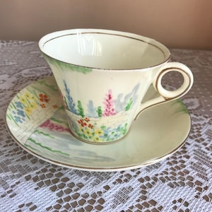 Op de afbeelding: Een vintage theekopje en schotelset met een bloemenmotief. Het kopje en de schotel zijn wit met een gouden rand en hebben een geschilderd tafereel van een tuin met bloemen en een bakstenen muur.