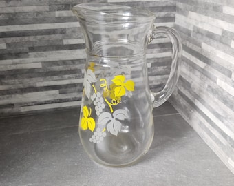 May include: A clear glass pitcher with a vine and grape design. The grapes are white and the leaves are yellow.