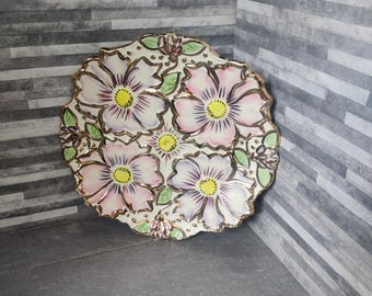 May include: A decorative ceramic plate featuring a floral design. The plate has a white base with pink, purple, and yellow flowers. The edges are trimmed with gold, and green leaves are interspersed among the flowers. The plate is round.