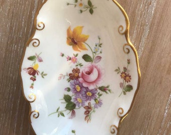 May include: A white oval-shaped porcelain dish with a gold rim and floral design. The dish features a bouquet of pink, yellow, and purple flowers with green leaves.