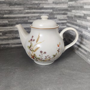 Può includere: Una teiera in ceramica bianca con un design floreale che presenta grano, bacche e foglie. La teiera ha un bordo marrone e un coperchio.