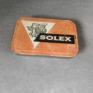 May include: Orange and white tin box with a black and white illustration of a carburettor and the word "SOLEX" in black letters.