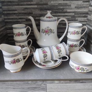 Puede incluir: Un juego de té de porcelana blanca con adornos florales y azules. El juego incluye una tetera, una jarra de crema, un azucarero, tazas y platillos. El diseño floral presenta flores rosas, moradas y azules.