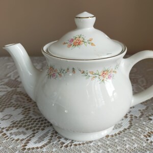 Puede incluir: Una tetera de porcelana blanca con tapa, asa y boquilla. La tetera está decorada con motivos florales en tonos rosa, naranja y verde. El borde y el asa tienen ribetes dorados. La tetera está sobre un mantel de encaje blanco.