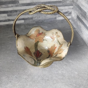 May include: A vintage decorative basket with a floral design. The basket has a cream interior with orange and red flowers. The handle is made of twisted gold-coloured metal.