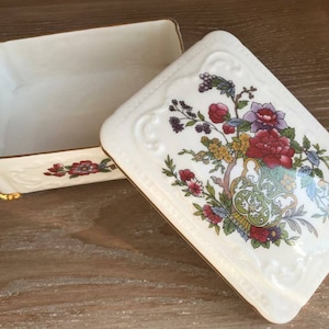 Puede incluir: Una caja de porcelana blanca con un borde dorado y un diseño floral. La tapa presenta un patrón floral con flores rojas, amarillas y verdes.