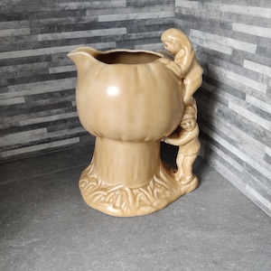 Op de afbeelding: Een keramische kan in de vorm van een paddenstoel met twee figuren die erop klimmen. De kan is lichtbruin van kleur en heeft een getextureerd oppervlak.