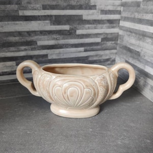 Pode incluir: Um vaso de cerâmica de cor creme com duas alças e um design decorativo de formas de leque sobrepostas. O vaso está sobre uma superfície cinza.