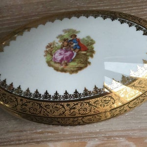 May include: Oval porcelain trinket box with a gold trim and a painted scene of a couple in a garden.