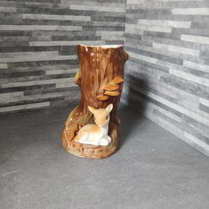 Puede incluir: Un jarrón de cerámica con forma de tocón de árbol, con un cervatillo en la base. El jarrón es marrón con detalles beige y blancos, y tiene una superficie texturizada. El cervatillo es marrón claro y blanco.