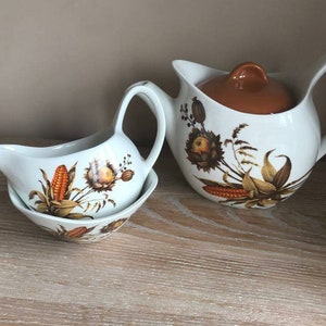 Könnte beinhalten: Ein weißes Keramik-Soßenboot und Sahne-Set mit einem Herbst-Ernte-Design. Das Set enthält ein Soßenboot mit passender Untertasse und einen Sahnekännchen mit Deckel. Das Design zeigt Mais, Weizen und andere herbstliche Elemente.