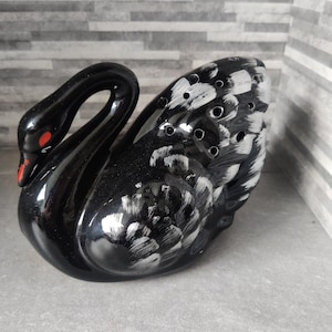 Könnte beinhalten: Glänzende schwarze Schwanfigur mit rotem Schnabel und Augen. Die Flügel des Schwans sind mit weißen Pinselstrichen und kleinen kreisförmigen Öffnungen verziert. Der Keramikvogel steht auf einer grauen Oberfläche.