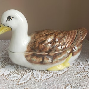 May include: A ceramic duck-shaped container. The head is white with a yellow beak, and the body is brown and white with a textured pattern. It has yellow feet and is likely a decorative item or serving dish.