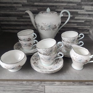 May include: A vintage tea set featuring a white teapot, cups, saucers, a sugar bowl, and a creamer. The set has a floral pattern in shades of blue and gray, with light blue accents on the handles and rims. The tea set is arranged on a gray surface.