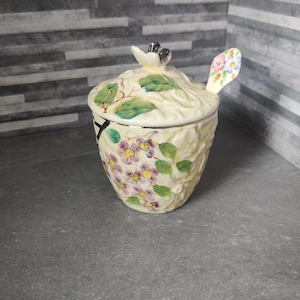 Könnte beinhalten: Ein cremefarbenes Keramikgefäß mit Deckel und Löffel. Das Gefäß ist mit lila Blumen und grünen Blättern verziert. Der Deckel hat ein Blumenmuster und einen kleinen Griff. Der Löffel hat ein Blumenmuster auf der Schaufel. Ein dekorativer Küchenartikel.