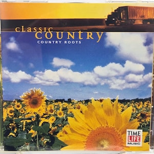 May include: A Classic Country Country Roots music CD in a clear plastic jewel case. The cover art features a field of yellow sunflowers under a blue sky with white clouds and a semi-truck silhouette. Text includes Time Life Music and Greatest Hits.