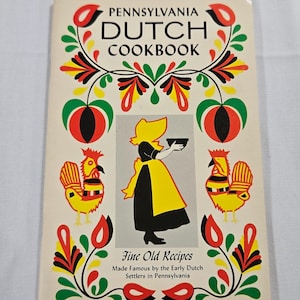 May include: A vintage cookbook titled "Pennsylvania Dutch Cookbook" with a cream-colored cover. The cover features colorful floral and rooster illustrations, along with a central image of a woman in traditional attire. The text reads "Fine Old Recipes".
