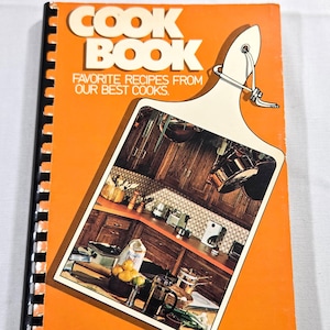May include: A vintage cookbook with an orange cover and spiral binding. The title "COOK BOOK" is in large white letters. A cutting board illustration features a kitchen scene. The text reads "FAVORITE RECIPES FROM OUR BEST COOKS."