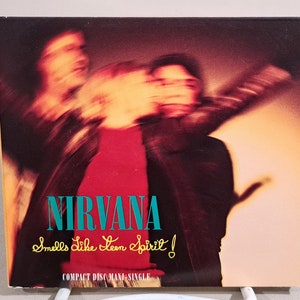 May include: Compact disc single for Nirvana's "Smells Like Teen Spirit." The album cover features a blurred image of three figures in front of a red shirt. The band's name is in teal, and the song title is in yellow.