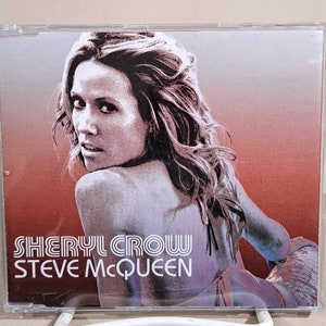 May include: A CD case with a photo of Sheryl Crow on the front. The background is a gradient of red and orange. The text "SHERYL CROW" and "STEVE MCQUEEN" is printed in black and white.