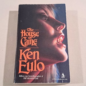 May include: A paperback book cover with a woman's face on the front. The woman has blonde hair and is wearing red lipstick. The title of the book is "The House of Caine" by Ken Fulo. The book is a million-copy bestselling author of "The Bloodstone".